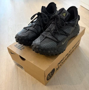 Nike ACG Mountain Fly Low sneakers - Svarta Nike ACG Mountain Fly Low sneakers med robust sula och reflekterande detaljer. Skorna har ett tekniskt meshmaterial, snörning och diskret Nike-logga på sidan. Perfekta för dig som gillar outdoor-vibe och streetstyle.