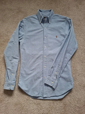 Ljusblå skjorta från Polo Ralph Lauren - Klassisk ljusblå skjorta från Polo Ralph Lauren i slim fit med button-down krage och broderad logga på bröstet. Tillverkad i mjuk bomull och har långa ärmar samt vita knappar framtill. Perfekt för en clean och stilren look.