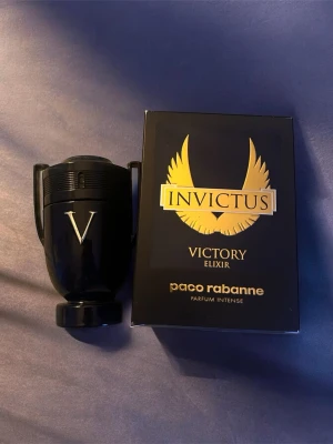 Paco Rabanne Invictus Victory Elixir - Exklusiv parfym från Paco Rabanne, Invictus Victory Elixir. Flaskan är svart med guldiga detaljer och har en unik pokalform. Förpackningen är svart med guldtext och en stor vinge, vilket ger en lyxig känsla. Parfymen är en intensiv doft för dig som vill sticka ut. Runt 90ml kvar i flaskan.