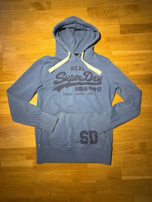 Superdry hoodie - Snygg blå hoodie från Superdry med stor logga och japansk text på bröstet. Storlek M och har inga defekter. Hör av dig vid frågor! Skickar snabbt vid snabb affär!