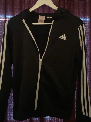 Svart Adidas hoodie med dragkedja - Svart hoodie från Adidas med vit dragkedja och klassiska vita ränder längs ärmarna. Adidas-logga på bröstet och huva. Tillverkad i mjukt material som passar perfekt för en sportig och avslappnad stil.