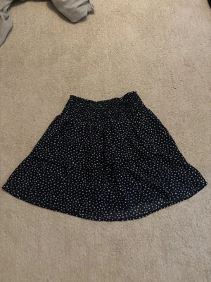 Svart kortkjol med vita prickar - Söt svart kortkjol med vita prickar och resår i midjan. Kjolen har en lätt utställd form och är gjord i ett tunt, mjukt material som ger ett flowy intryck. Perfekt för dig som gillar en enkel men ändå trendig stil.