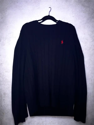 Svart stickad tröja från Ralph Lauren - Svart stickad tröja från Ralph Lauren med klassisk röd logga broderad på bröstet. Tröjan har rund halsringning och långa ärmar, perfekt för lager-på-lager under kyliga dagar. Snygg kabelstickad struktur som ger en tidlös look.