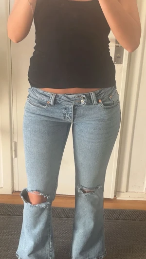 Blåa jeans med hål - Nya! Säljer ett par ljusblå bootcut jeans med slitna hål på båda knäna. Jeansen har låg midja och klassisk femficksdesign. Perfekta för en avslappnad och trendig look. Materialet är denim i bomull.