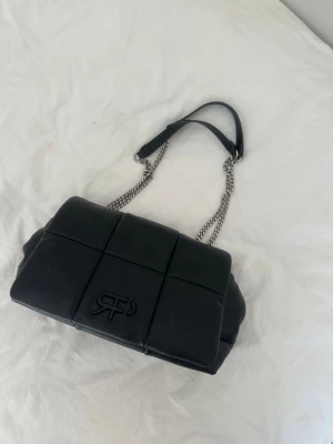 Svart quiltad handväska från River Island - Snygg svart quiltad handväska från River Island med kedjerem och vadderad design. Väskan har ett stort rutmönster och logga på framsidan. Perfekt accessoar för att lyfta din outfit.