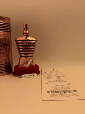 Jean Paul Gaultier Le Male Elixir - Jean Paul Gaultier Le Male Elixir parfym i en unik, kroppformad flaska i metallic roséguld. Flaskan har horisontella ränder och kommer med en matchande cylinderformad metallförpackning och röd sammetsbas. Snygg och lyxig design som sticker ut i badrumshyllan.