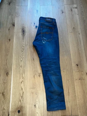 Blå slitna jeans från Nudie Jeans - Säljer ett par blå jeans från Nudie Jeans med slitningar och distressed detaljer på framsidan. Klassisk femficksmodell med normal midja och bra passform. Jeansen är gjorda i denim och har en snygg tvättad look som passar till det mesta.