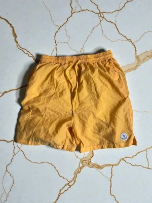 Gula badshorts från Moncler - Snygga gula badshorts från Moncler med elastisk midja och snörning framtill. Shortsen har en bakficka med knapp och Moncler-logga på vänster ben. Perfekta för strand eller pool under soliga dagar.