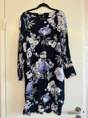 Blommig svart långklänning med dragkedja - Svart långklänning med stora blommor i lila, vitt och grått. Klänningen har långa ärmar med dragkedjor vid ärmsluten och en lös, bekväm passform. Tillverkad i ett mjukt, lätt material som känns skönt mot huden.