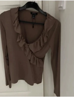 Brun volangblus från H&M - Snygg brun blus  med långa ärmar och volangdetaljer längs den djupa v-ringningen. Tillverkad i ett mjukt och lätt material som sopar fint. Perfekt för fester/speciella stunder. 