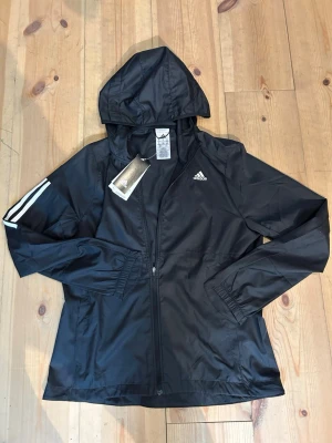 Svart vindjacka med huva från Adidas - Snygg svart vindjacka från Adidas med huva och dragkedja framtill. Jackan har vita ränder på ena ärmen och Adidas-logga på bröstet. Tillverkad i lätt och tåligt material, perfekt för blåsiga dagar. Elastiska muddar vid ärmsluten för extra komfort.