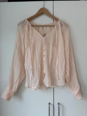 Ljusrosa blus med knappar - Säljer en ljusrosa blus med v-ringning och knappar framtill. Blusen har långärmade, löst sittande ärmar med smock vid ärmsluten. Tillverkad i ett tunt, skirt tyg som ger en luftig känsla. Lappen är kvar.