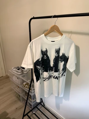 Vit Stüssy t-shirt med tryck - Vit t-shirt från Stüssy med ett stort svart grafiskt tryck framtill som föreställer tre hundar och Stüssy-loggan. Klassisk passform med rund hals och korta ärmar. Perfekt för dig som gillar streetwear och vill sticka ut.