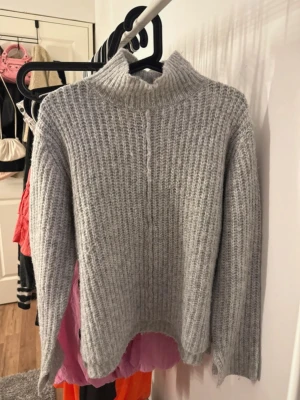 Ljusgrå stickad polotröja - Mysig ljusgrå stickad tröja med hög krage och lång ärm. Tröjan har ett grovt ribbstickat mönster och är tillverkad i ett mjukt, fluffigt material som känns skönt mot huden. 