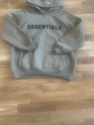 Size S. Beige Essentials hoodie med tryck. Aldrig andvänt  - Säljer en beige hoodie från Essentials med stor svart logga på bröstet och diskret text på ärmen. Hoodien har huva och magficka, samt ribbade muddar vid ärmar och nederkant. Perfekt för en avslappnad och trendig look.