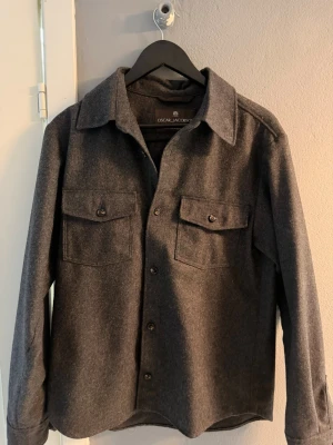 Mörkgrå overshirt från Oscar Jacobson - Stilren mörkgrå overshirt från Oscar Jacobson i mjukt material. Skjortjackan har klassisk krage, två bröstfickor med lock och knappar framtill. Perfekt lager på lager-plagg med avslappnad passform och tidlös design. Nypris 2499kr. Använd 2 gånger. Storlek M. Pris kan diskuteras vid snabb affär.