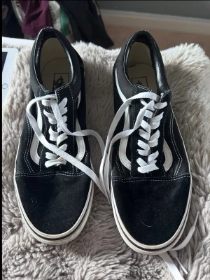 Svarta och vita Vans Old Skool sneakers - Klassiska Vans Old Skool sneakers i svart canvas och mocka med vita detaljer och vit sula. Skorna har den ikoniska vita sidoremsan och vita skosnören. Perfekta för en streetwear-look och passar till det mesta.