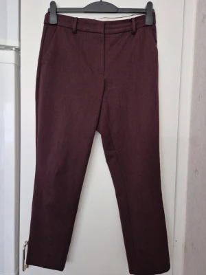 Kostymbyxor - Snygga mörkröda/bruna kostymbyxor med raka ben och klassisk passform. Byxorna har hällor för skärp, diskreta sidofickor och en bakficka. Perfekta för en stilren look. Materialet ser ut att vara en mjuk och lite stretchig blandning. Finn gul fläck.