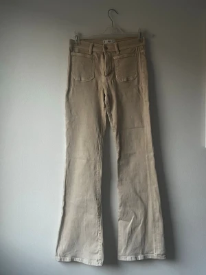 Beiga utsvängda byxor med medelhög midja Manchester  - Snygga beiga jeans med utsvängda ben och hög midja. Jeansen har klassiska fem fickor och raka sömmar. Liten defekt se bild!
