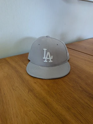 Grå LA-keps från New Era - Snygg grå keps från New Era med LA-logga broderad framtill i vitt. Klassisk flat peak  baksida. Mjuk Perfekt för dig som gillar streetwear och vill ha en stilren accessoar till din outfit.