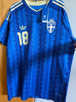 Blå Sverige fotbollströja Adidas - Kom gärna med prisförslag om ni inte gillar priset!Säljer en blå Sverige fotbollströja från Adidas med retroinspirerat mönster, nummer 18 och namnet Ayari på ryggen. Tröjan har vita och gula detaljer, broderad landslagssköld och Adidas-logga. Tillverkad i lätt och ventilerande material, perfekt för match eller träning.