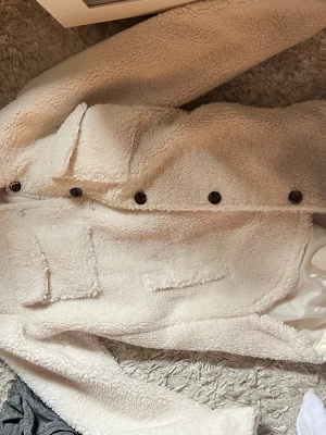 Vit teddyjacka från Lindex - Mysig vit teddyjacka från Lindex med stora fickor framtill och knappar längs hela framsidan. Jackan har en fluffig och mjuk yta i teddy material. Perfekt för uppklädnad även när det är kallt och jacka till en mysig höstdag