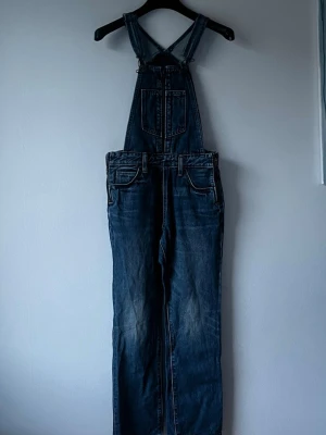 Mörkblå denim hängselbyxa Levis  - Säljer en klassisk mörkblå denim hängselbyxa med raka ben och justerbara axelband. Byxdressen har flera fickor fram och bak samt detaljerade sömmar. 