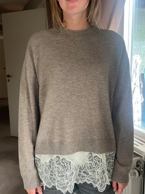 Beige stickad tröja med spetskant - Säljer en beige stickad tröja med rund hals och långa ärmar. Tröjan har en unik detalj med vit spetskant nedtill som ger en snygg lager-på-lager-look. Perfekt för dig som gillar en avslappnad men ändå stilren outfit.