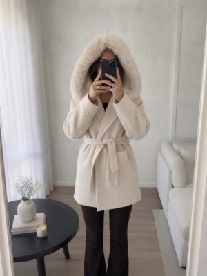 Beige kappa med pälsdetaljer - Elegant beige kappa med stor huva och pälsdetaljer. Kappan har ett knytbälte i midjan och praktiska fickor framtill. Perfekt för kyliga dagar när du vill hålla stilen. 