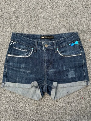 Vintage Levi's jeansshorts med diamantdetaljer - Snygga vintage mörkblå jeansshorts från Levi's med uppvikt benkant och glittriga strassdetaljer på fickorna!💋Så coola! Klassisk femficksmodell med knapp och dragkedja. Perfekta för sommaren och ger en cool vibe till din outfit!! Shortsen är lågmidjade/mid rise💞Sydda och dekorerade av mig med noggrannhet, passion & kärlek❤️Hör av vid frågor, pris är förhandlingsbart. Mått: 34,5cm (midja), 6cm (innerben), 22cm (grenhöjd)🤍