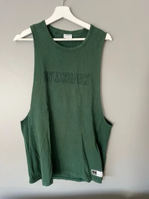 Grön ärmlös linnetopp från Gymshark - Mörkgrön linnetopp från Gymshark med broderad logga framtill. Toppen är ärmlös med djupa ärmhål och har en avslappnad passform, perfekt för träning. Tillverkad i mjuk bomull för maximal komfort.