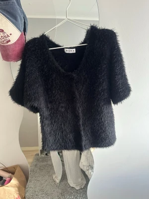 Svart fluffig kofta från Wera - Säljer en svart, kortärmad och fluffig kofta från Wera. Koftan har en djup v-ringning och är gjord i ett mjukt, lurvigt material som ger en trendig look. Perfekt att styla över ett linne eller t-shirt för en cool outfit.