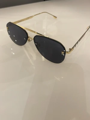 Svarta aviator solglasögon med gulddetaljer - Snygga solglasögon i klassisk aviator-modell med svarta glas och tunna guldfärgade bågar. Glasen är mörka och har små dekorativa nitar samt en diskret logga på ena linsen. Perfekta för en trendig look i solen.