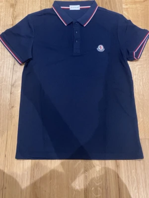 Marinblå pikétröja från Moncler - Snygg marinblå pikétröja från Moncler med klassisk krage och knappar framtill. Tröjan har vita och röda detaljer på krage och ärmslut samt Moncler-logga på bröstet. Perfekt för en stilren och sportig look.