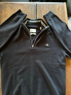 Svart halfzip tröja GANT - Svart stickad halfzip tröja från GANT i mjuk lammull. Tröjan har halv dragkedja, ribbad krage med vita ränder och broderad logga på bröstet. Perfekt för dig som gillar klassiskt och stilrent