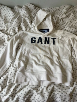 Vit hoodie från GANT med tryck - Säljer en vit hoodie från GANT med stor svart logga på bröstet. Hoodien har huva och en klassisk magficka framtill. Tillverkad i mjukt bomullsmaterial och har ribbade muddar vid ärmslut och nederkant. Perfekt för en avslappnad och stilren look.