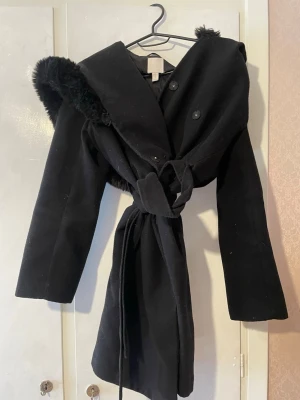 Svart kappa med stor huva och knytskärp - Snygg svart kappa med stor fluffig huva och knytskärp i midjan. Kappan har två knappar framtill och en mjuk, pälsliknande kant på huvan. Perfekt för kalla dagar och ger en cool siluett. Passar dig som vill ha en stilren men ändå statement ytterjacka.