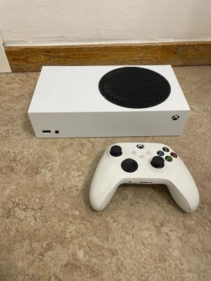 Xbox Series S med vit handkontroll - Säljer en Xbox Series S i stilren vit färg tillsammans med en matchande vit trådlös handkontroll. Konsolen har en kompakt, rektangulär design med en stor svart cirkulär ventil på ovansidan. Perfekt för gaming och digitala spel.