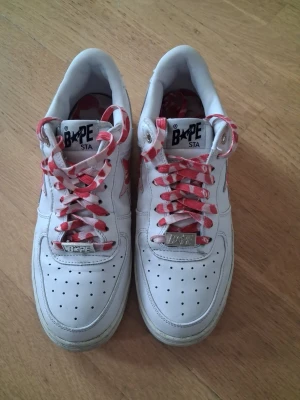 Vita BAPE STA sneakers med röda detaljer - Säljer ett par vita BAPE STA sneakers med röda stjärndetaljer på sidan och röda/vita mönstrade skosnören. Skorna har perforerad tåbox och klassisk låg siluett. Tillverkade i läder med tydliga streetwear-vibbar och ikonisk branding på plösen. (Lådan är lite trasig)