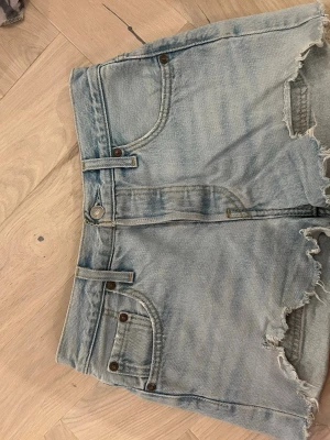 Ljusblå Levi's jeansshorts hög midja - Säljer ett par klassiska ljusblå jeansshorts från Levi's med hög midja och råa, fransiga benslut. Shortsen har fem fickor, bälteshällor och Levi's ikoniska läderpatch bak. Perfekta för sommaren och riktigt snygga till sneakers eller sandaler.