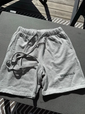 Grå Essentials Fear of God shorts - Säljer ett par grå shorts från Essentials Fear of God med elastisk midja och långa snören framtill. Shortsen har fickor på sidorna och en bakficka. Perfekta för chill dagar och har en clean look med logga på benet.