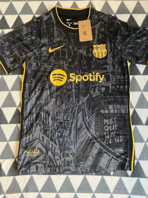 Barcelona svart matchtröja Nike M - Säljer en svart Barcelona matchtröja från Nike med gula detaljer och tryck. Tröjan har klubbmärke, Spotify-logga och texten 'MES QUE UN CLUB' på framsidan. Unik grafisk design inspirerad av stadens arkitektur. Perfekt för fotbollsfans!