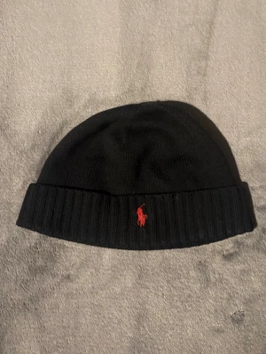 Polo ralph Lauren mössa  - Snygg svart ribbstickad mössa från Polo Ralph Lauren med uppvikt kant och klassisk röd logga broderad framtill. Perfekt accessoar för kyliga dagar och enkel att matcha med olika outfits.