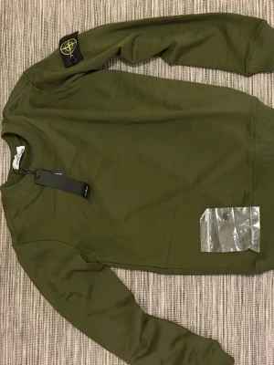 Olivgrön sweatshirt från Stone Island - Olivgrön sweatshirt från Stone Island med klassisk rund halsringning och ribbade muddar. Tröjan har den ikoniska Stone Island-loggan på vänster ärm och kommer med originaltaggar. Perfekt för en avslappnad och stilren look.