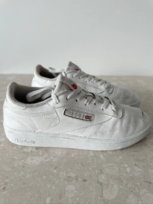 Vita Reebok Club C 85 sneakers storlek 39  - Klassiska vita Reebok Club C 85 sneakers i läder med lågt skaft och snygga detaljer som logga och brittiska flaggan på sidan. Skorna har vit sula och diskret Reebok-märke på hälen. Perfekta för en clean och stilren look.