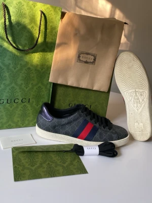 Gucci sneakers med blå och röda ränder - Snygga Gucci sneakers i svart canvas med GG-mönster, blå och röda sidoremsor samt vita sulor. Skorna har svarta skosnören och detaljer i mörkblått läder vid hälen. Klassisk låg modell med exklusiv känsla och tydlig Gucci-logga på sulan.
