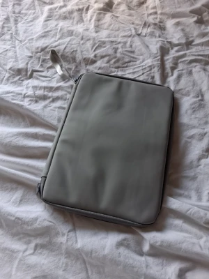 Ljusgrå laptopväska med dragkedja - Snygg och stilren ljusgrå datorväska med dragkedja och praktiska innerfack. Väskan har en slät utsida och insidan har både meshficka och vanliga fack för tillbehör. Perfekt för att hålla koll på laddare och småprylar.