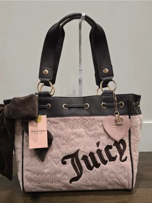 Juicy Couture daydreamer tote  - Trendig handväska från Juicy Couture i beige sammet med broderad logga och svarta detaljer. Väskan har breda svarta handtag, guldfärgade metalldetaljer och ett hjärtformat hänge med krona. Perfekt för dig som gillar statement-accessoarer.