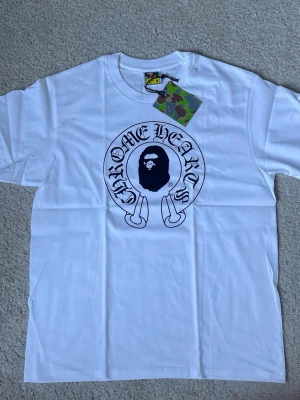 Vit bape T-shirt med Chrome Hearts tryck - Vit t-shirt med stort svart Chrome Hearts-tryck och ikoniskt ansikte på bröstet. Klassisk passform och rund hals. T-shirten har korta ärmar och är tillverkad i mjuk bomull. Perfekt för en streetwear-look.