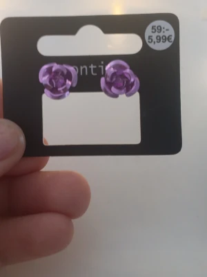 Lila rosformade örhängen  - Snygga örhängen från montini i form av lila rosor. De är små och har en metallisk finish som ger ett coolt intryck. Perfekta för dig som vill ha något unikt och färgglatt till din stil. Stiftet är i metall och örhängena är lätta att bära.